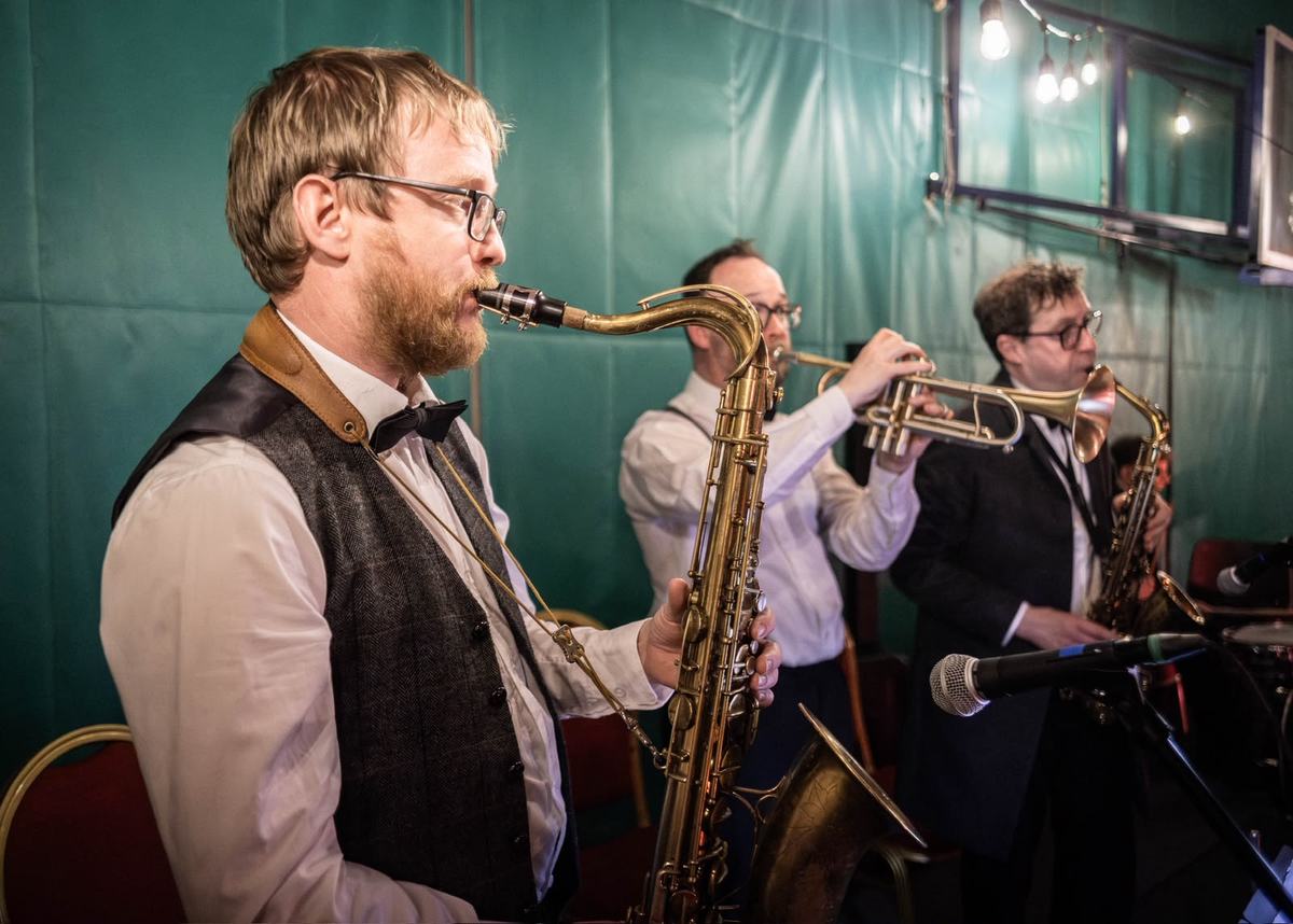 A saxophone section in waistcoats and bow ties, playing in front of a green curtain.