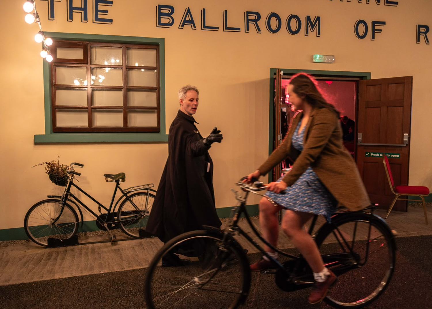 Outside a country hall painted with the words The Ballroom of Romance, a woman cycling past and a caped figure standing in the doorway.