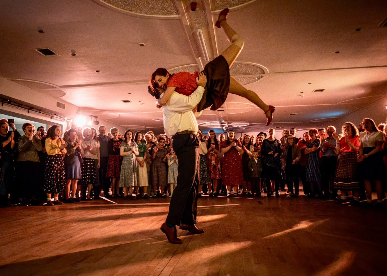 A dancer lifted high overhead on a packed dance hall floor, the crowd ringed around them under warm light.